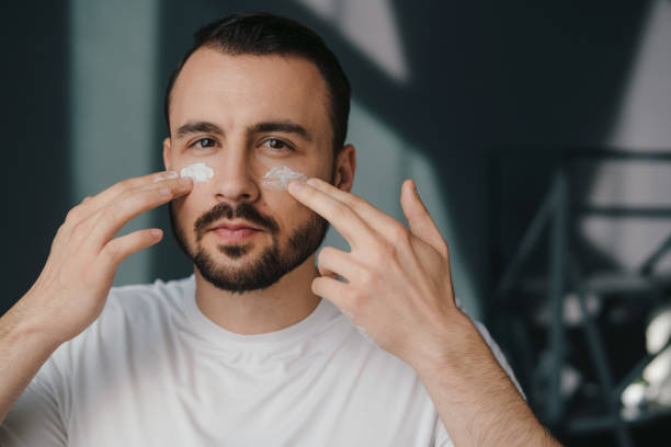 istock Handsome unshaven young man looking at camera applying moisturizing face cream on his cheeks. Portrait of man performing daily self-care. Beauty routine indoors. Beauty treatment. Positive person. Beauty face. Facial skincare. Health care. 1480062407