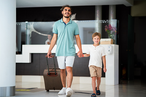 Happy father and son holding hands and walking in hotel lobby whit luggage