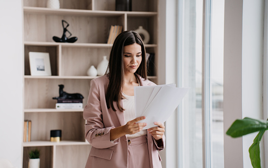 Brunette Asian businesswoman in suit holds sheet of papers looks at documents stands at window against bookshelves. Successful lawyer explores issue at office. Financial bill.