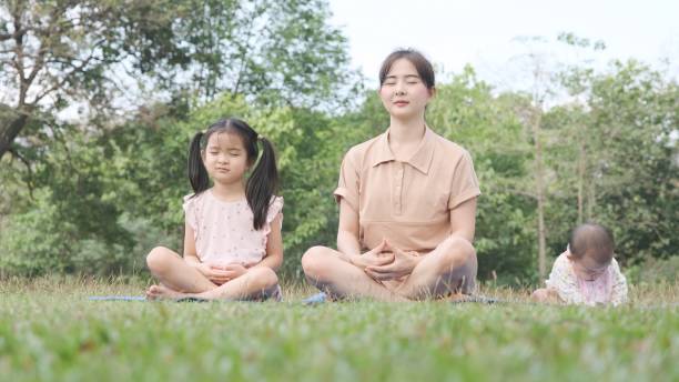 asiatische junge mutter und tochter, die in yoga-asanas sitzen, posieren mit einem entzückenden kleinen mädchen, das frech auf matten-yoga im park spielt. - yoga good posture women little girls stock-fotos und bilder