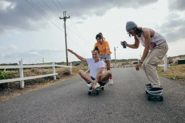 Photo of Three Asian good friends are skateboarding together outdoors, and they are using smartphones to record their activity and upload it to social media.