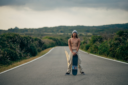An Asian young man is walking on an outdoor road, holding a skateboard and ready for a skateboarding experience.