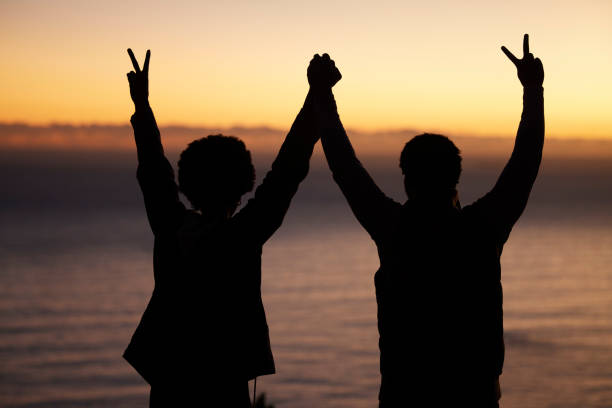 pôr do sol, praia e silhueta de celebração do casal, sucesso ou vitória e de mãos dadas na sombra, noite ou escuridão. céu, oceano e pessoas vencedoras na retaguarda com sinal de paz para o mar, caminhadas ou metas de viagem - placard holding celebration women - fotografias e filmes do acervo