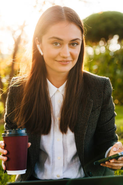 portrait of a young adult caucasian business woman using mobile phone in city park, drinking coffee cup while looking at camera. sitting woman outdoors. people using laptop. - surfing wireless vertical outdoors lifestyles imagens e fotografias de stock