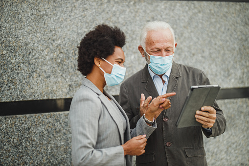 Shot of successful senior businessman and his black female colleague with protective mask using digital tablet and having a discussion while standing against a wall of corporate building during COVID-19 pandemic.