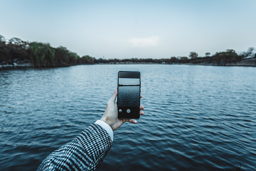 A man is outdoors using his phone to shoot a lake at sunset