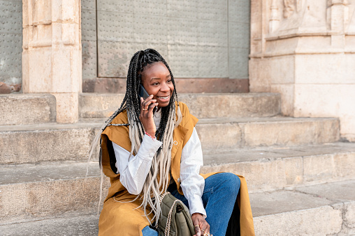 smiling african american woman with braids sitting on the stairs in the city talking on the cellphone