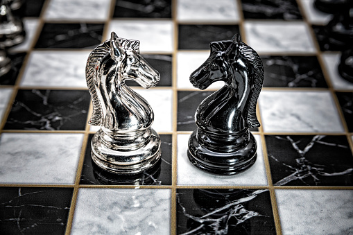Two opposing knights positioned in front of each other on a fully set up chess board, shot in studio suggesting a metaphor for confrontation or battle.