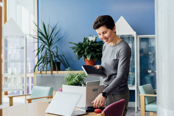 istock Satisfied newcomer middle aged neutral gender female company worker standing in shared coworking office unpacking box with personal belongings at new workplace having first working day concept. 1480102695