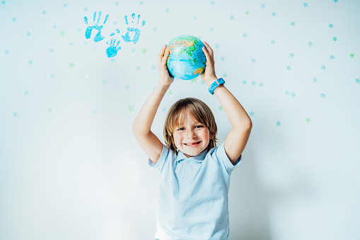 Smiling caucasian boy holding an earth globe model over his head on the background with stars and family hand prints on the white wall. Save the planet, Earth Day. Global peace and unity concept