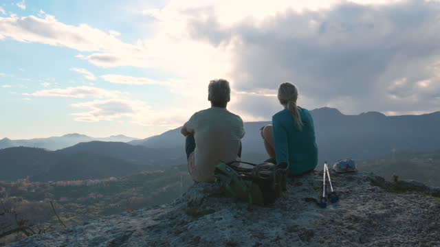 Mature hiking couple relax on summit viewpoint above distant mountains