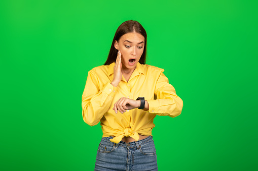 Deadline. Shocked Millennial Lady Looking At Smartwatch On Hand In Shock Standing Posing Over Green Studio Background. Omg, I'm Late, Time is Tickling Concept