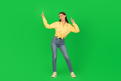 Full Length Shot Of Joyful Woman Dancing Listening To Music With Earbuds Earphones Posing In Studio Over Green Background. Great Song, Musical Application Advertisement