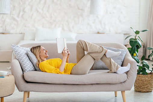 Happy pretty young blond european woman in yellow clothes lies on sofa and reads book in minimalist living room interior, profile. Hobby, literature at weekend free time during covid-19 quarantine