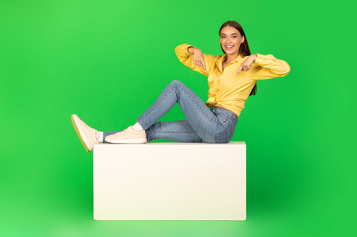 Look There. Happy Female Pointing Fingers Down At White Stand Smiling To Camera Sitting Posing Over Green Studio Background. Great Offer, Text Advertisement Concept