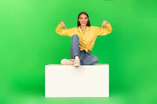 Great Offer. Excited Woman Pointing Fingers Down Sitting On White Stand Advertising Something Posing Smiling To Camera Over Green Background In Studio. Look There Concept