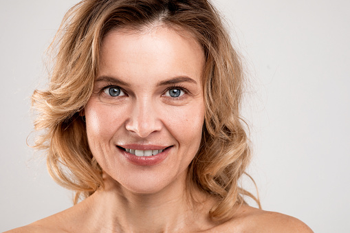 Closeup Portrait Of Smiling Attractive Middle Aged Woman With Bare Shoulders Posing Over Light Studio Background, Happy Mature Lady With Beautiful Aged Skin Looking At Camera, Copy Space
