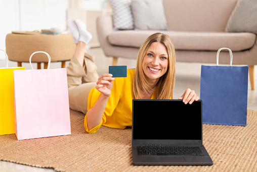 Smiling millennial caucasian lady shopaholic with many packages from store use credit card show laptop with blank screen, enjoy online shopping in living room interior. Website, sale, cashback at home