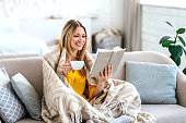 Smiling pretty young european blonde lady wrapped in plaid drinks favorite hot drink and reads book on sofa