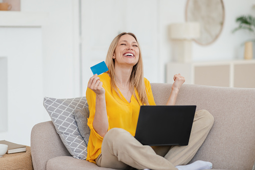 Happy excited young blond european female making victory gesture with credit card and laptop, rejoices to successful deal on sofa in living room interior. Shopaholic at home during covid-19 quarantine
