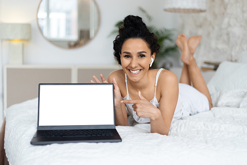 Smiling arab millennial female woke up, lies on bed in wireless headphones, points finger at laptop with blank screen in bedroom interior. Good morning at home with device, blogger recommendation