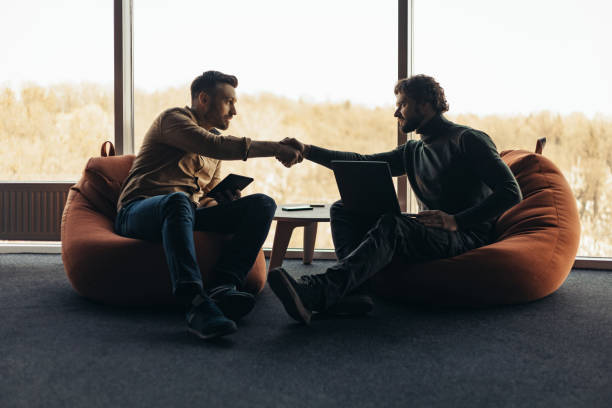 istock Male colleagues with portable computers shaking hands, making business agreement, sitting in bean bag chairs at office 1480107737