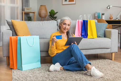Smiling european old woman with gray hair uses credit card and tablet for online shopping in room interior with packages with purchases. Big sale for shopaholic at home, delivery during covid-19