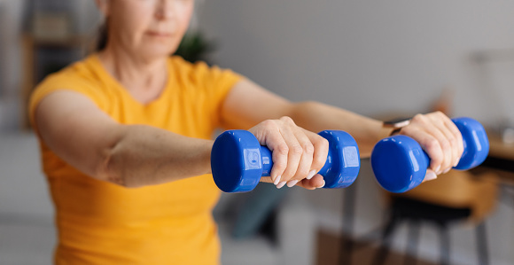 Sporty aged woman lifting dumbells up, training in living room interior, working on arms strength, selective focus, panorama. Senior female doing dumbbell workout at home