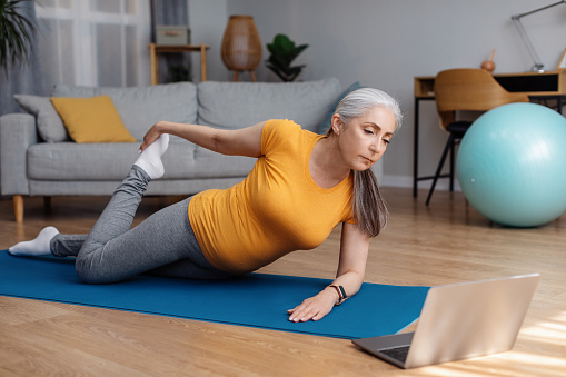 Domestic training. Active senior woman doing legs quad stretching exercises while watching video tutorial on laptop, looking at screen. Sport, online gym concept
