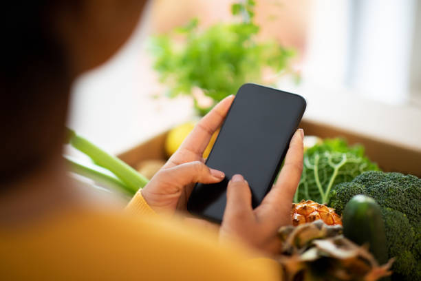 mujer afroamericana millennial desempaca caja de cartón con verduras orgánicas usa smartphone con pantalla en blanco - equipment housework remote domestic kitchen fotografías e imágenes de stock
