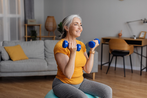 Happy senior woman exercising with dumbbells at home, sitting on fitball and strengthening her arm muscles, having domestic strength training, free space