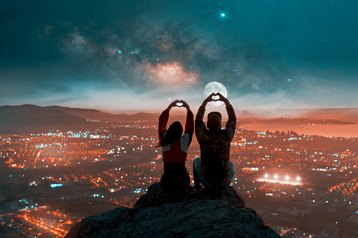 silhouette of a heterosexual couple sitting on top of a mountain making a silhouetted heart shape sign with their hands against the background of moon and stars over the city