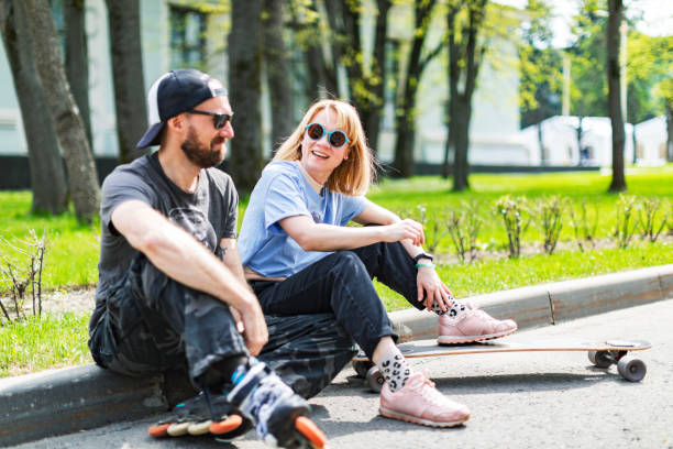 Adult hipster couple with longboard and roller skate sitting in park stock photo
