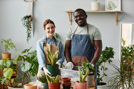 Waist up portrait of two smiling gardeners looking at camera indoors while posing with lush green plants