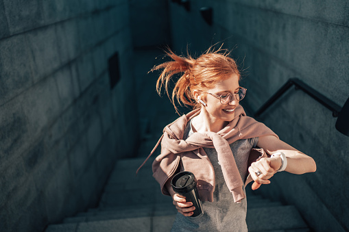 Beautiful skinny redhead woman enjoying fitness exercising surrounded with concrete walls in city urban environment. She is also listening to music with wireless earbuds.