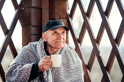 Elderly man wrapped in warm plaid drinks hot coffee or tea from white mug while sitting in wooden gazebo near cottage outdoor.