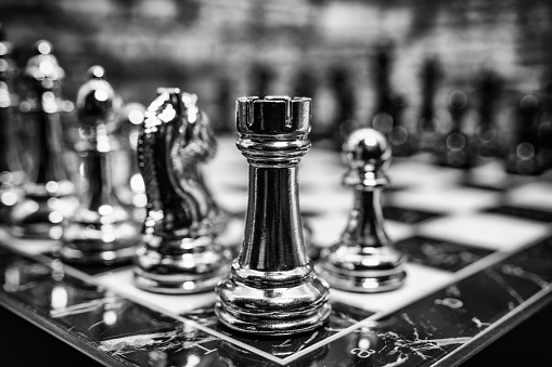 A fully set up chess board with both the black and white pieces set in their squares with the white pawn making its opening move shot in studio against a brick background.