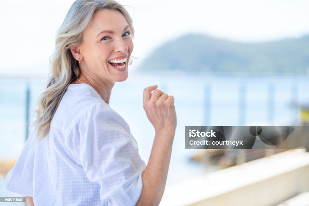 Beautiful mature woman portrait at waterfront house. Beautiful mature woman portrait at waterfront house.  She is happy and smiling with the sea behind her. She looks relaxed and could be on vacation. Women Stock Photo