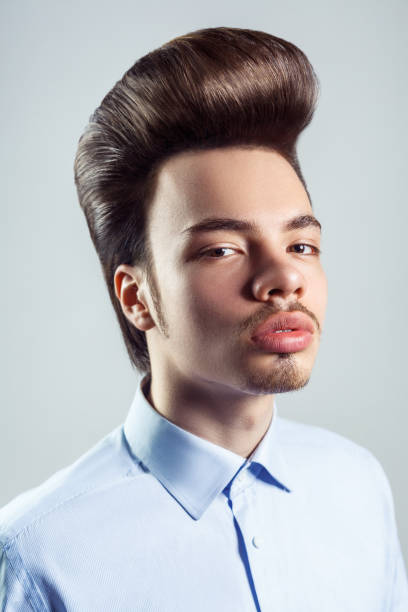 Slelf-confident man with mustache with retro pompadour hairstyle, looking with serious expression. Portrait of young adult self-confident man with beard and mustache with retro classic pompadour hairstyle, looking at camera with serious expression. Indoor studio shot isolated on gray background. rockabilly hair men stock pictures, royalty-free photos & images