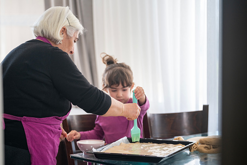 Candid photo of senior woman with white hair and 4,5 years old granddaughter preparing pastry for cooking in domestic kitchen. Shot indoor under daylight.