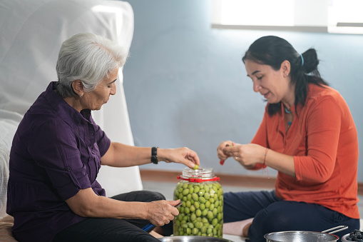 Candid photo of senior woman and mature adult friend sitting on the floor in living room and preparing green olives for seasoning. Window is in view. Shot indoor under daylight.