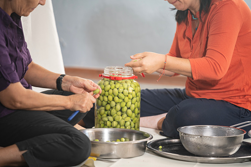 Candid photo of senior woman and mature adult friend sitting on the floor in living room and preparing green olives for seasoning. Window is in view. Shot indoor under daylight.