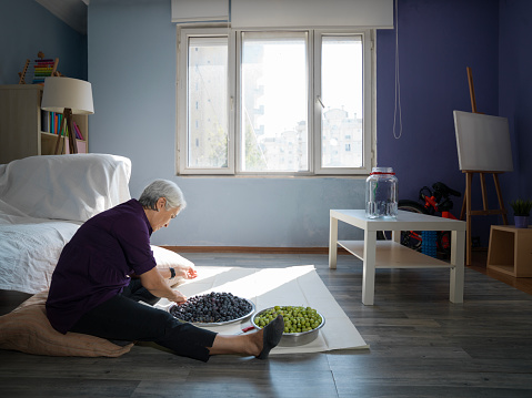Candid photo of senior woman sitting on the floor in living room and preparing green olives for seasoning. Window is in view. Shot indoor under daylight with a medium format camera.