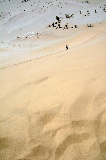 A man climbs the sand from a small oasis to a hill in the desert along a path. View from a distance from a drone