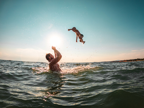 Happy kid playing with his father in the sea on a fresh summer afternoon. The little boy is flying in the air and jumping into the water. They are on summer vacations.