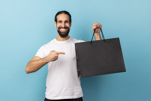 Portrait of optimistic man with beard wearing white T-shirt pointing at shopping bags, looking at camera with pleased happy facial expression. Indoor studio shot isolated on blue background.