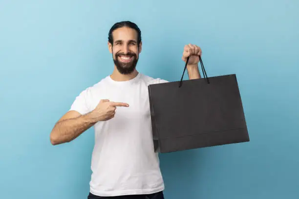 Photo of Man pointing at shopping bags, looking at camera with pleased happy facial expression.