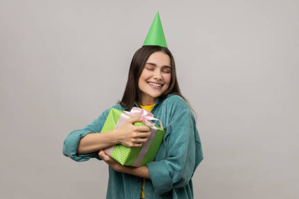 счастливая женщина обнимает подарок и улыбается с выражением удовольствия, обнимается с любовью в подарок на день рождения. - hat conical стоковые фото и изображения