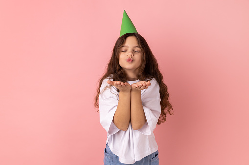 Portrait of little girl wearing white T-shirt with party cone on head sending air kiss, sharing love, amorous feelings on Valentines day. Indoor studio shot isolated on pink background.