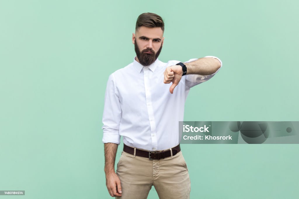 Businessman criticizing bad quality with thumbs down displeased grimace, showing dislike gesture. Bearded businessman wearing shirt criticizing bad quality with thumbs down displeased grimace, showing dislike gesture, expressing disapproval. Indoor studio shot isolated on light green background. 20-24 Years Stock Photo